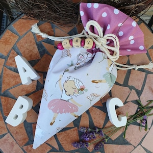 Könnte beinhalten: Ein Stoffbeutel mit Kordelzug und Ballerina-Aufdruck, mit rosa Tutu und floralen Akzenten. Der Beutel ist mit einer weißen Kordel und Holzperlen mit den Buchstaben A, B und C gebunden. Weiße Holzbuchstaben A, B und C sind ebenfalls vorhanden.