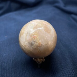 Op de afbeelding: Een grote, lichtroze bol van gepolijst selenietkristal op een metalen standaard. Het kristal heeft een doorschijnende, troebele uitstraling met zichtbare interne strepen.