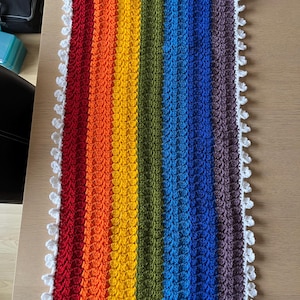 May include: A rectangular, handmade crochet blanket with a vibrant rainbow color scheme. The blanket features vertical stripes of red, orange, yellow, green, blue, and purple. A white, scalloped border frames the edges. The blanket is placed on a wooden surface.