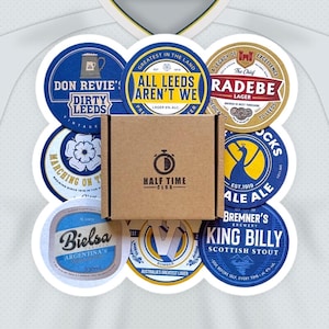 May include: A brown cardboard box with the text "Half Time Club" on the front, surrounded by nine round coasters with various designs and text. The coasters feature images of a football, a peacock, a beer mug, a flower, and a man's face. The text on the coasters includes "All Leeds Aren't We", "Don Revie's Dirty Leeds", "Radebe", "Marching On Together", "Bielsa", "King Billy", "Est. 1919", "Bomber", and "The Chief".