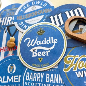 K&ouml;nnte beinhalten: Eine Sammlung von blauen und wei&szlig;en Untersetzern mit verschiedenen Designs. Die Untersetzer zeigen den Text "The Owls", "Hillsborough", "Sin", "Waddle Beer", "Magic Chris", "Barry Bann", "Scottish Sto", "Herid", "1989-1995", "Probably the best midfielder in the world", "79-92" und "Ho".