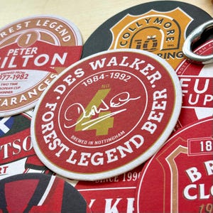 May include: A collection of red, white, and gold coasters featuring the names of Nottingham Forest Football Club legends. The coasters feature the names "Peter  ilton", "Des Walker", and "Collymore".