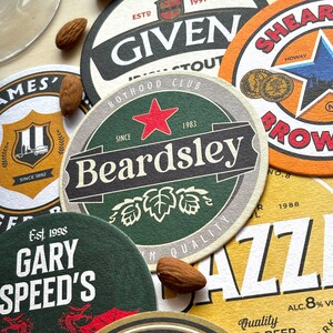 May include: A collection of colorful round coasters with various designs and text. The coasters feature a green and brown coaster with the text "Beardsley" and "Since 1983", a yellow and orange coaster with the text "Shear Brow" and "Howay", a green coaster with the text "Gary Speed's" and "Est. 1998", a brown and white coaster with the text "Given" and "Boyhood Club", and a yellow coaster with the text "Quality" and "ALC. 8% Vol."