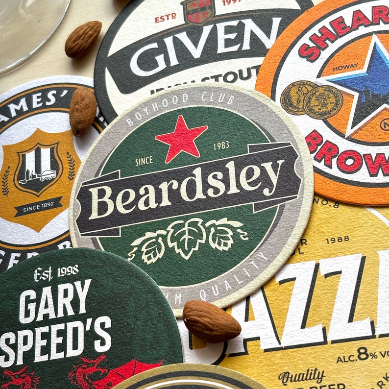 May include: A collection of colorful round coasters with various designs and text. The coasters feature a green and brown coaster with the text "Beardsley" and "Since 1983", a yellow and orange coaster with the text "Shear Brow" and "Howay", a green coaster with the text "Gary Speed's" and "Est. 1998", a brown and white coaster with the text "Given" and "Boyhood Club", and a yellow coaster with the text "Quality" and "ALC. 8% Vol."