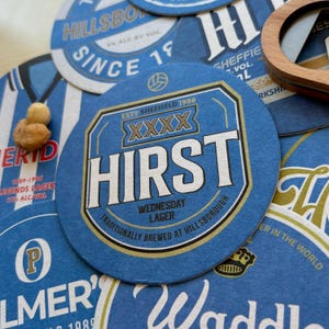 K&ouml;nnte beinhalten: Blaue und goldene runde Untersetzer mit dem Text "Hirst" und "Wednesday Lager" in wei&szlig;er und goldener Schrift. Die Untersetzer zeigen auch den Text "Traditionally brewed at Hillsborough" und "Est. Sheffield 1986" in kleinerer Schrift.