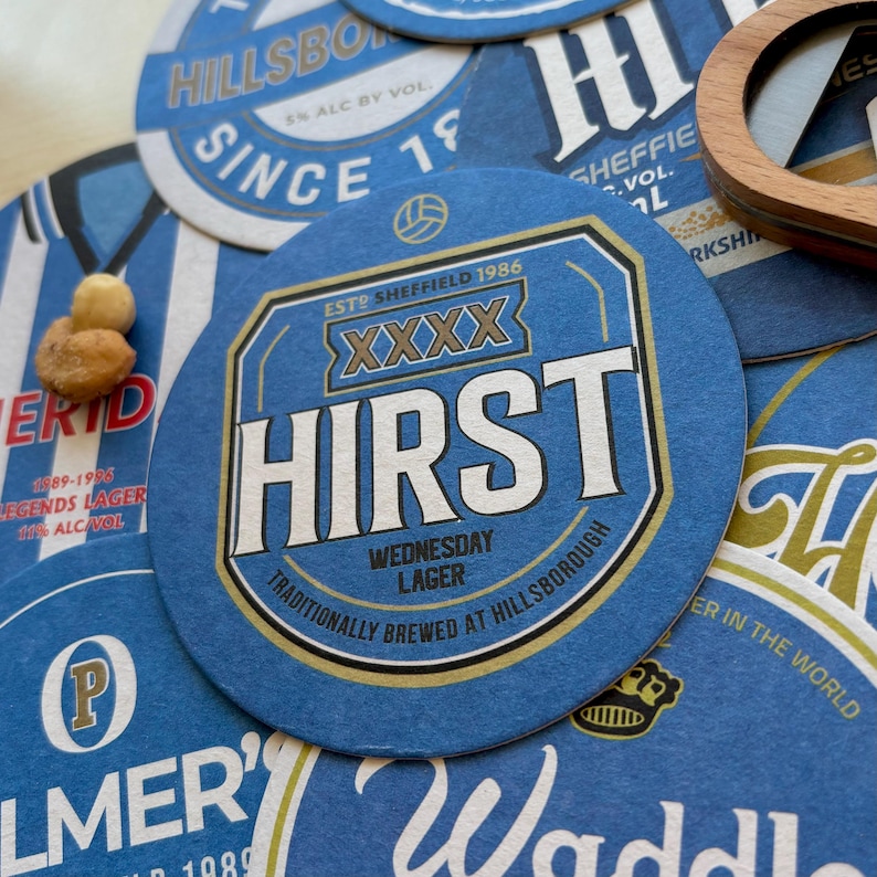 K&ouml;nnte beinhalten: Blaue und goldene runde Untersetzer mit dem Text "Hirst" und "Wednesday Lager" in wei&szlig;er und goldener Schrift. Die Untersetzer zeigen auch den Text "Traditionally brewed at Hillsborough" und "Est. Sheffield 1986" in kleinerer Schrift.