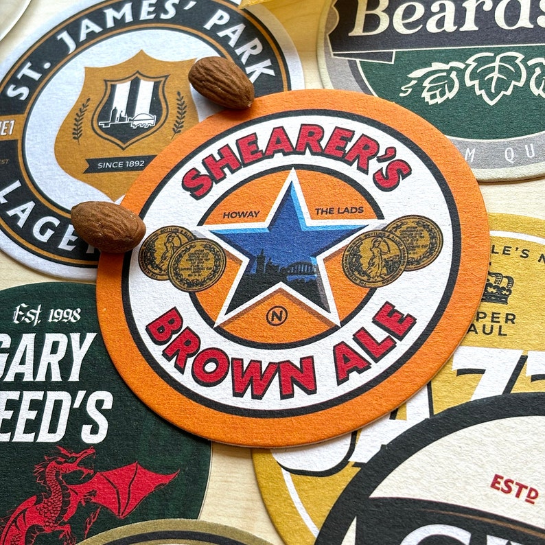 May include: A collection of colorful round coasters with various designs. One coaster features a star with the words "Shearer's Brown Ale" and "Howay The Lads". Another coaster has a crest with the words "St. James' Park" and "Since 1892".