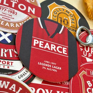 May include: A red and black beer coaster with a football jersey design. The jersey has the name "PEARCE" and the text "1985-1997 LEGENDS LAGER 3% ALC/VOL".