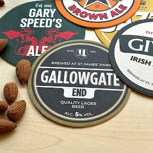 May include: A collection of six different beer coasters. The coasters are round and feature various designs, including a dragon, a brewery logo, and a pub name. The coasters are made of cardboard and have a brown, black, and white color scheme.