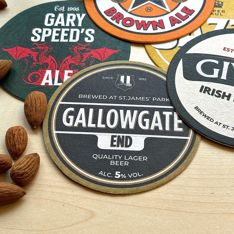 May include: A collection of six different beer coasters. The coasters are round and feature various designs, including a dragon, a brewery logo, and a pub name. The coasters are made of cardboard and have a brown, black, and white color scheme.