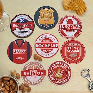 May include: Ten round coasters with red, white, and black designs featuring names and numbers of famous soccer players. The coasters are on a wooden surface with almonds in a bowl.