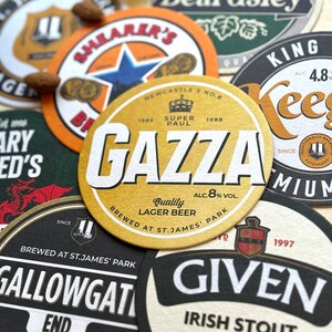 May include: A collection of round, colorful coasters with various brewery logos and names. The coasters are made of cardboard and have a textured surface. The coasters feature the names "Shearer's", "Gazza", "Gallowgate", "Keegan's", and "Given".