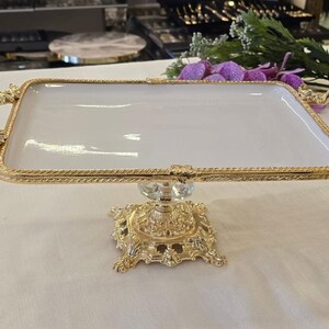 May include: A white rectangular serving tray with a gold-coloured, ornate frame and handles. The tray is supported by a gold-coloured, decorative base. The handles have a floral design. The tray is likely used for serving food or displaying items.