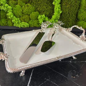 Könnte beinhalten: Ein silberfarbenes Kuchenservierset auf einem weißen rechteckigen Tablett. Das Set enthält ein Kuchenmesser und einen Servierer, beide mit verzierten Griffen. Das Tablett hat dekorative Griffe und einen detaillierten silbernen Rand.