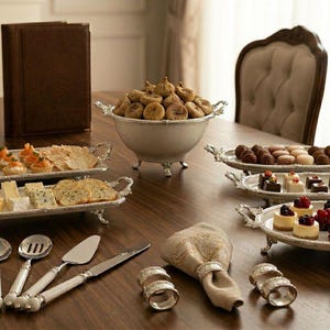 May include: A table set for a formal occasion, with silver trays laden with appetizers and desserts. Includes a bowl of figs, pastries, cheese, and serving utensils. A brown leather-bound book and decorative napkin rings complete the elegant arrangement.