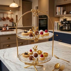 May include: A two-tiered gold-colored serving tray with glass plates, filled with small pastries. The tray has a decorative bow at the top and ornate details. Gold forks and spoons are placed on the marble countertop.