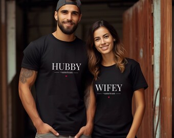 Może przedstawiać: Dwa czarne t-shirty z białym napisem "HUBBY" i "WIFEY". Oba mają "#sameteam" i czerwone serce. Zestaw pasujących koszulek dla par.