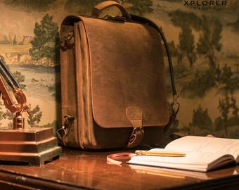 Könnte beinhalten: Eine braune Ledertasche mit Klappenverschluss, Griff und verstellbarem Schulterriemen. Die Tasche hat messingfarbene Hardware und ist mit "XPLORER CRAFTS" geprägt. Die Tasche liegt auf einem Holzschreibtisch mit Notizbuch und Stift.