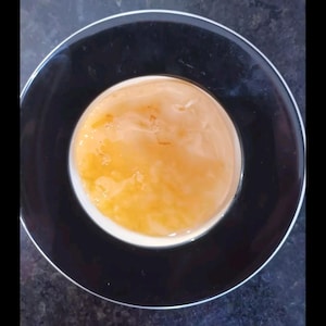 May include: A black plate with a white interior contains a light orange, semi-transparent liquid. The liquid has a slightly textured surface. The plate's rim is black, creating a contrast with the lighter centre.