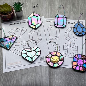 May include: A set of eight iridescent stained glass gemstone ornaments, each with a different shape, including a heart, diamond, square, and circle. The ornaments are hanging from chains and are displayed on a white sheet of paper with numbered grids.