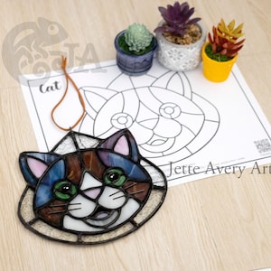 Può includere: Un acchiappasole in vetro colorato a forma di gatto con pezzi di vetro marrone, blu, verde e bianco. Il gatto ha un'espressione allegra e guarda a destra. L'acchiappasole è appeso a un cordoncino marrone. L'immagine include anche una pagina da colorare in bianco e nero di un gatto con il testo "Jette Avery Art" in basso.