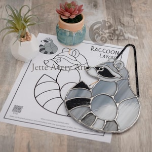 May include: A stained glass raccoon suncatcher with a black chain. The raccoon is grey with black accents and is in a curled up position. The suncatcher is on a white sheet of paper with a black and white outline of a raccoon and the text "Raccoon Layout" and "Jette Avery Art".