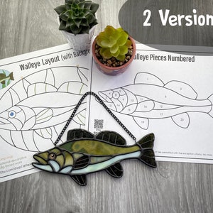 Puede incluir: Un pez lucioperca de vitral con piezas de vidrio verde, marrón y blanco. El pez está colgado de una cadena y se encuentra sobre una superficie de madera. Hay dos hojas de papel con un contorno en blanco y negro del pez, una con las piezas numeradas y otra con un fondo.