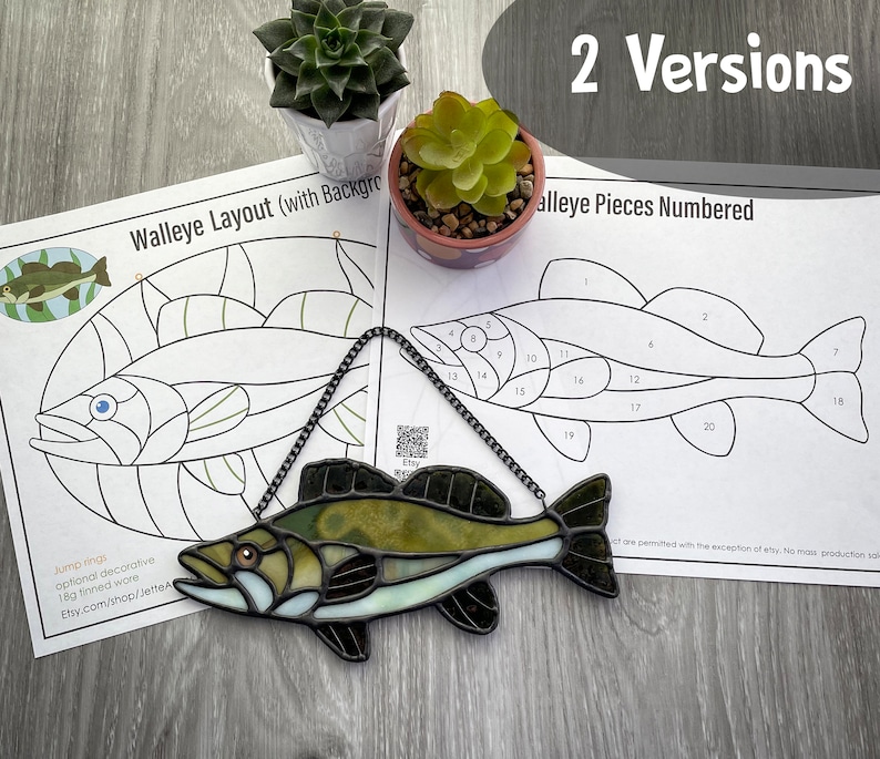 May include: A stained glass walleye fish with green, brown and white glass pieces. The fish is hanging from a chain and is on a wooden surface. There are two sheets of paper with a black and white outline of the fish, one with the pieces numbered and one with a background.