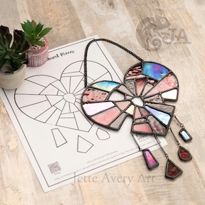 May include: A heart-shaped stained glass suncatcher with a black metal frame. The heart features pink, red, and iridescent glass. The suncatcher hangs from a chain and has dangling glass pieces. A paper with a heart design is in the background.