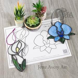 May include: Stained glass flower ornaments, a white calla lily and a blue orchid, sit on a paper with flower outlines. The calla lily has a yellow center and green leaves. The orchid has blue petals and green leaves. Small potted succulents are in the background.