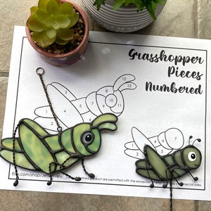 May include: A green stained glass grasshopper with black wire legs and a hanging chain. The grasshopper is numbered with pieces 1 through 13. The image also includes a black and white outline of a grasshopper with the text "Grasshopper Pieces Numbered".