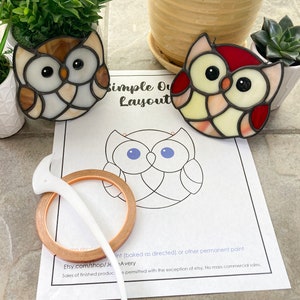 Op de afbeelding: Twee gebrandschilderde uil-versieringen, een met bruin en wit glas en de andere met rood en wit glas, worden getoond op een wit vel papier met een zwart-witte omtrek van een uil. Het papier bevat ook de tekst "Simple Owl Layout" en instructies voor het gebruik van de omtrek.