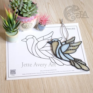 Peut inclure: Ornement d'oiseau en vitrail bleu clair, beige et transparent, suspendu par une ficelle argentée. Le motif est sur papier blanc avec "Jette Avery Art". De petites plantes en pot sont en arrière-plan. Le texte "Pieces" est également visible.