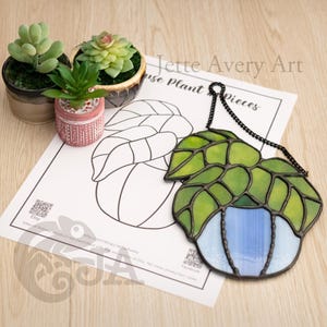 Op de afbeelding: Een hangend glas-in-loodwerk van een kamerplant met groene bladeren en een blauwe pot. De plant hangt aan een zwarte ketting. De afbeelding bevat ook een zwart-witte omtrek van de plant op een wit vel papier met de tekst "Jette Avery Art House Plant Pieces" bovenaan.