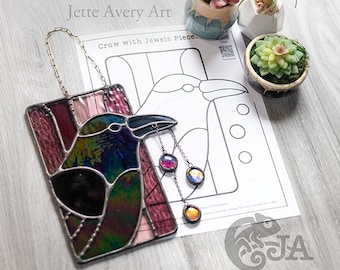 May include: Stained glass crow art piece with iridescent and jewel-toned glass. The crow has a black beak and is hanging from a chain. The artwork is rectangular and features a design of a crow with jewels. The text "Crow with Jewels Piece" is visible.