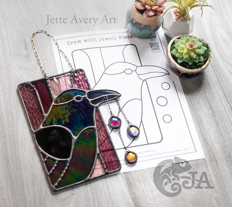 May include: Stained glass crow art piece with iridescent and jewel-toned glass. The crow has a black beak and is hanging from a chain. The artwork is rectangular and features a design of a crow with jewels. The text "Crow with Jewels Piece" is visible.