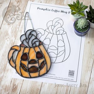 Könnte beinhalten: Ein Buntglas-Suncatcher in Form einer Kürbis-Kaffeetasse mit orangefarbenen Glaselementen, dunklen Lötlinien und einer silbernen Kette zum Aufhängen, platziert auf einer Papiervorlage mit der Aufschrift Pumpkin Coffee Mug Pattern.