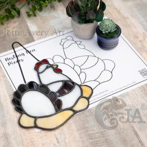 Puede incluir: Una gallina de nido de vidrio coloreado con detalles en negro, blanco, rojo y amarillo. La gallina está sentada en un nido con una cadena negra unida a la parte superior. La imagen incluye el texto "Nesting Hen Pieces".