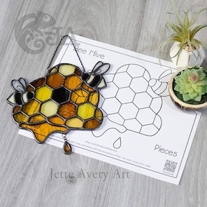 May include: Stained glass bee hive with yellow, amber, and black honeycomb design, two bees, and a hanging chain. The text "Bee Hive" is above the hive. A paper with a bee hive outline and the word "Pieces" is next to it.