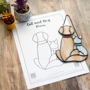 May include: A stained glass cat and dog suncatcher with a cream and brown dog and a blue cat. The suncatcher is hanging from a black chain. The image also includes a black and white outline of the cat and dog on a white sheet of paper with the text "Cat and Dog Pieces".