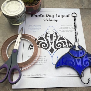 May include: A printed guide with instructions for creating a stained glass manta ray. The guide includes a black and white outline of the manta ray, along with text describing the process. A blue stained glass manta ray is shown hanging from a chain.