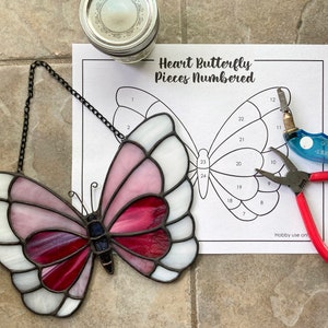Puede incluir: Una mariposa de vidrio emplomado con piezas de vidrio rosa, blanco y rojo. La mariposa está colgada de una cadena y está junto a un patrón impreso de una mariposa con piezas numeradas. El texto en el patrón dice "Heart Butterfly Pieces Numbered".