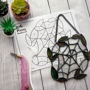 Puede incluir: Una telaraña de vidrio coloreado de metal negro con hojas verdes. La telaraña está numerada con una guía de patrones para ensamblar las piezas de vidrio coloreado.