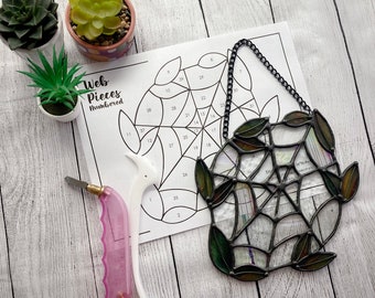 May include: A black metal stained glass spider web with green leaves. The web is numbered with a pattern guide for assembling the stained glass pieces.