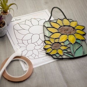 May include: A stained glass sunflower craft kit with a black metal frame and a printed paper template. The kit includes a roll of copper foil tape and a tool for applying the tape. The paper template has the words "Flower Pieces" printed on it.