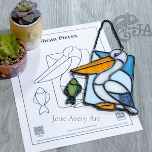 Puede incluir: Obra de arte de un pelícano de vidrieras con un pez en el pico, colgado de una cadena negra. El pelícano tiene vidrio blanco, azul y naranja. La obra de arte está sobre papel blanco con el texto "Pelican Pieces" y "Jette Avery Art".