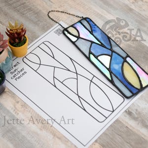 Pode incluir: Um apanhador de sol abstrato em vitral com uma moldura de metal preta, apresentando tons de azul, branco e amarelo. O apanhador de sol é retangular e pendurado por uma corrente. Um papel com um contorno de design e o texto "Abstract Suncatcher Pieces" está ao lado.