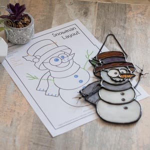 Könnte beinhalten: Ein Schneemann-Anhänger aus Buntglas mit einem braunen Hut, schwarzen Augen und einer Karottennase. Der Anhänger hängt an einer schwarzen Kette und befindet sich neben einem gedruckten Schneemann-Muster mit dem Text "Snowman Layout".