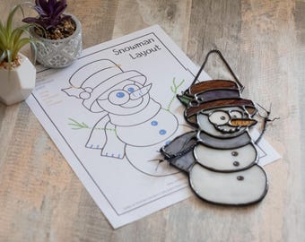 May include: A stained glass snowman ornament with a brown hat, black eyes, and a carrot nose. The ornament is hanging from a black chain and is next to a printed snowman pattern with the text "Snowman Layout".
