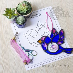 May include: A stained glass bird ornament in shades of blue and purple with a hanging crystal, placed on a paper pattern labeled Jette Avery Art. Includes a pink glass cutter, iridescent scissors, and two small potted succulent plants.